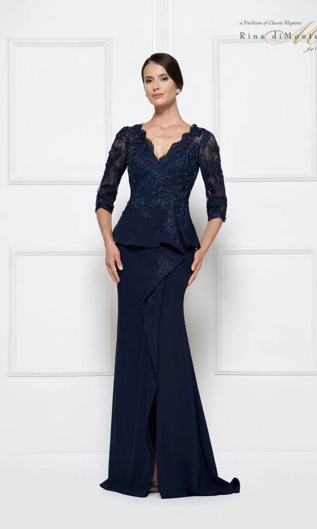 A woman stands before a white paneled wall in the Rina Di Montella RD2685, a navy long formal dress with lace sleeves, a scalloped neckline, and a fitted sheath skirt.